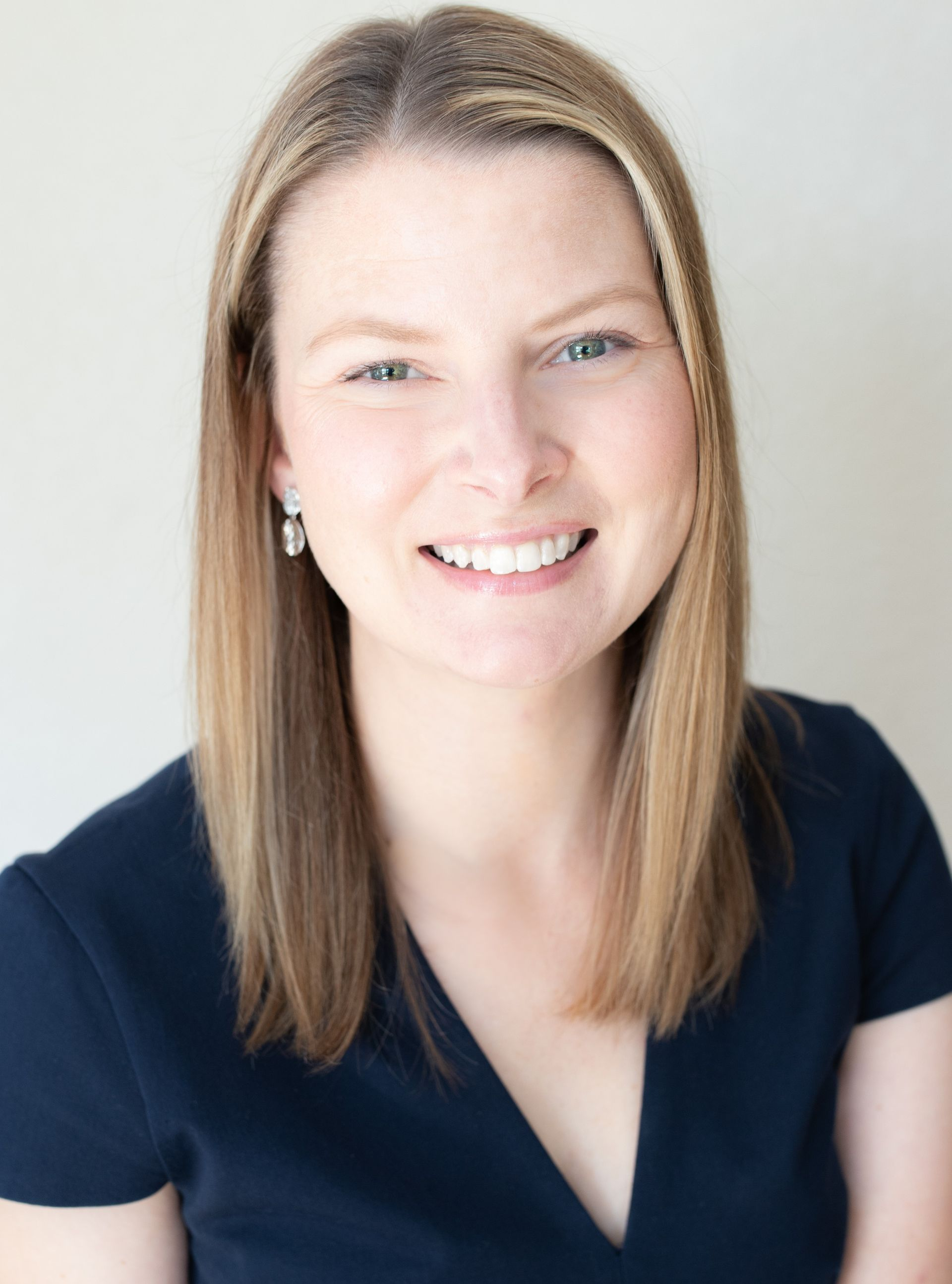 A smiling person with shoulder-length light brown hair wearing a dark blue top against a neutral background.