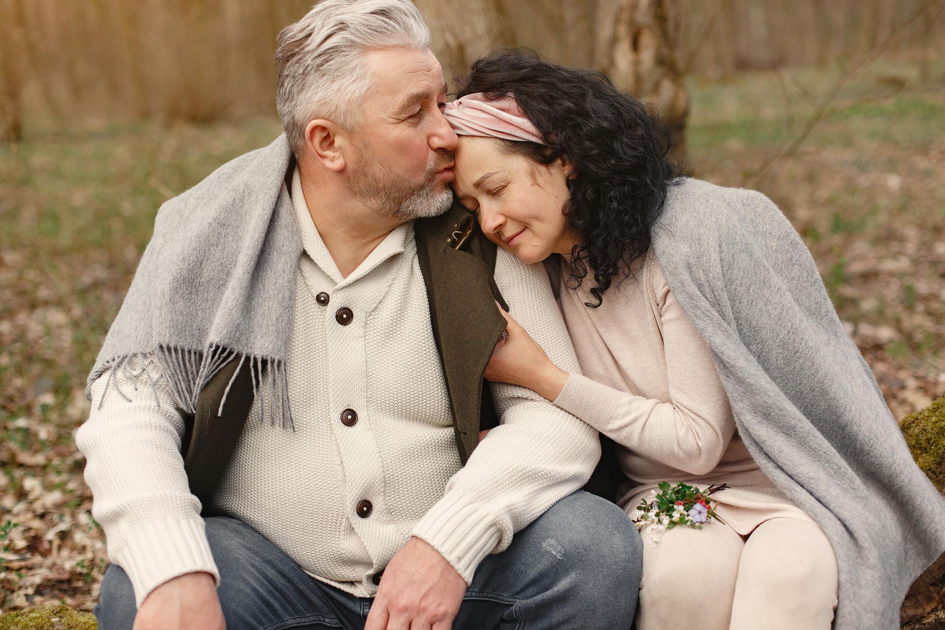 A couple wrapped in a grey blanket sitting in a wooded area, with one kissing the other's forehead.