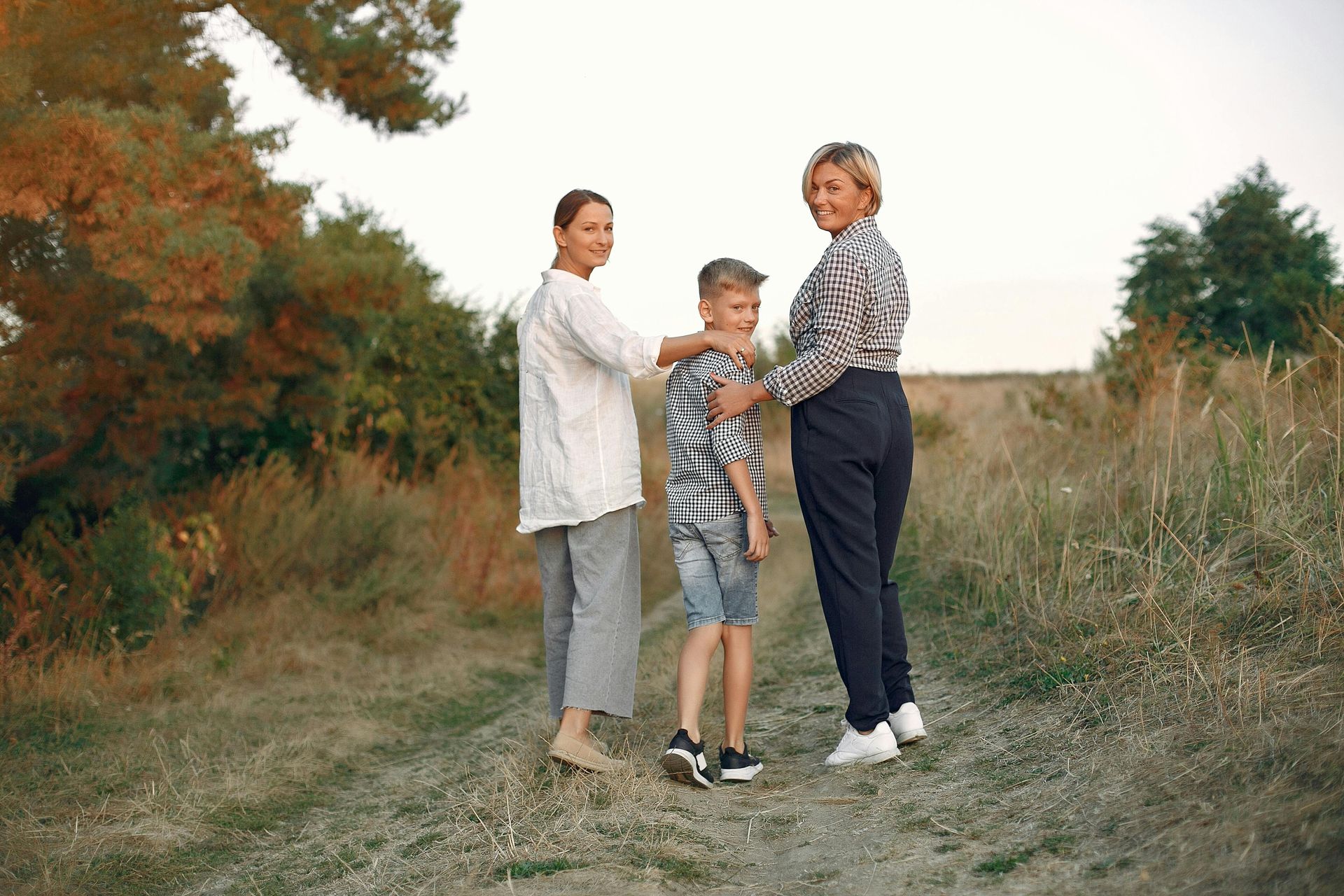 Three people walking together on a dirt path, turning back to look at the camera in a grassy, wooded area.