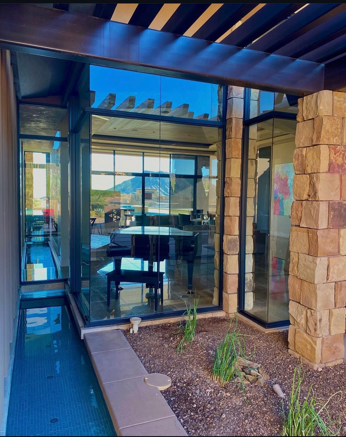 Exterior view with large windows, stone wall, water feature, and pergola. Mountains and interior visible through the glass.