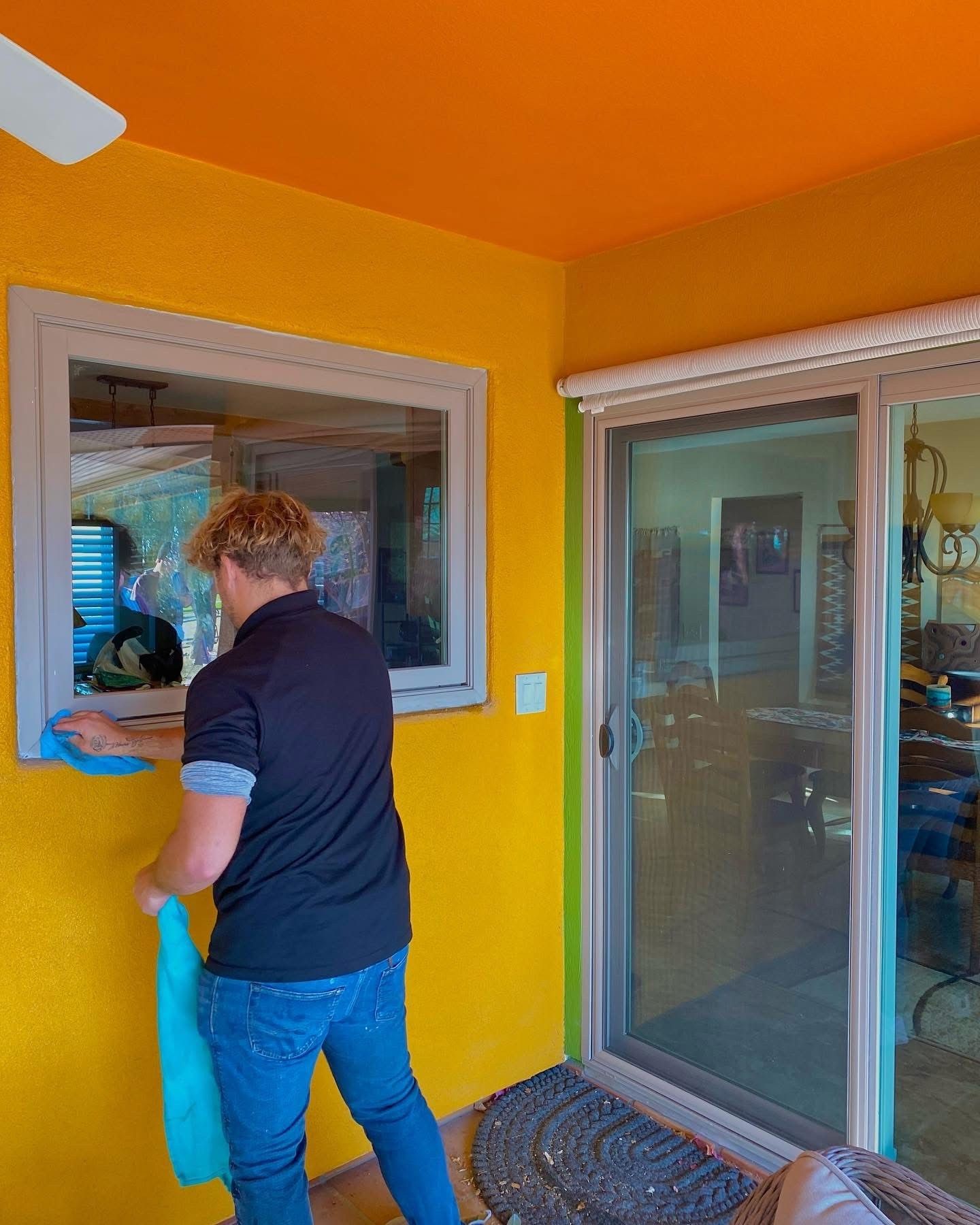 Person cleaning a window on a yellow exterior wall.  Bright orange ceiling, sliding glass door.