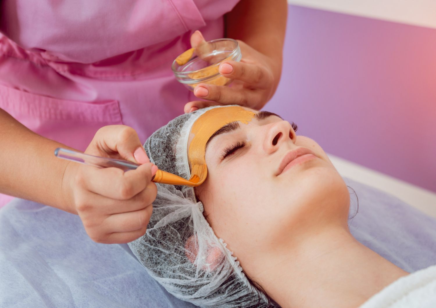 Person receiving a facial treatment; cosmetician applying orange mask with a brush.