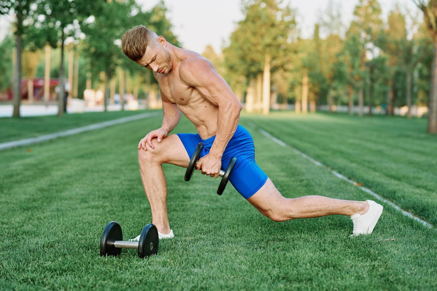 Man lunging with a dumbbell on green grass in a park. He is shirtless with blue shorts and white shoes.