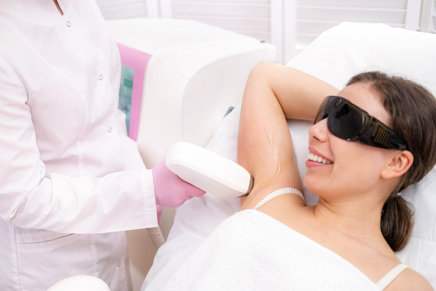 Woman getting laser hair removal on her armpit at a clinic, wearing protective glasses, smiling.
