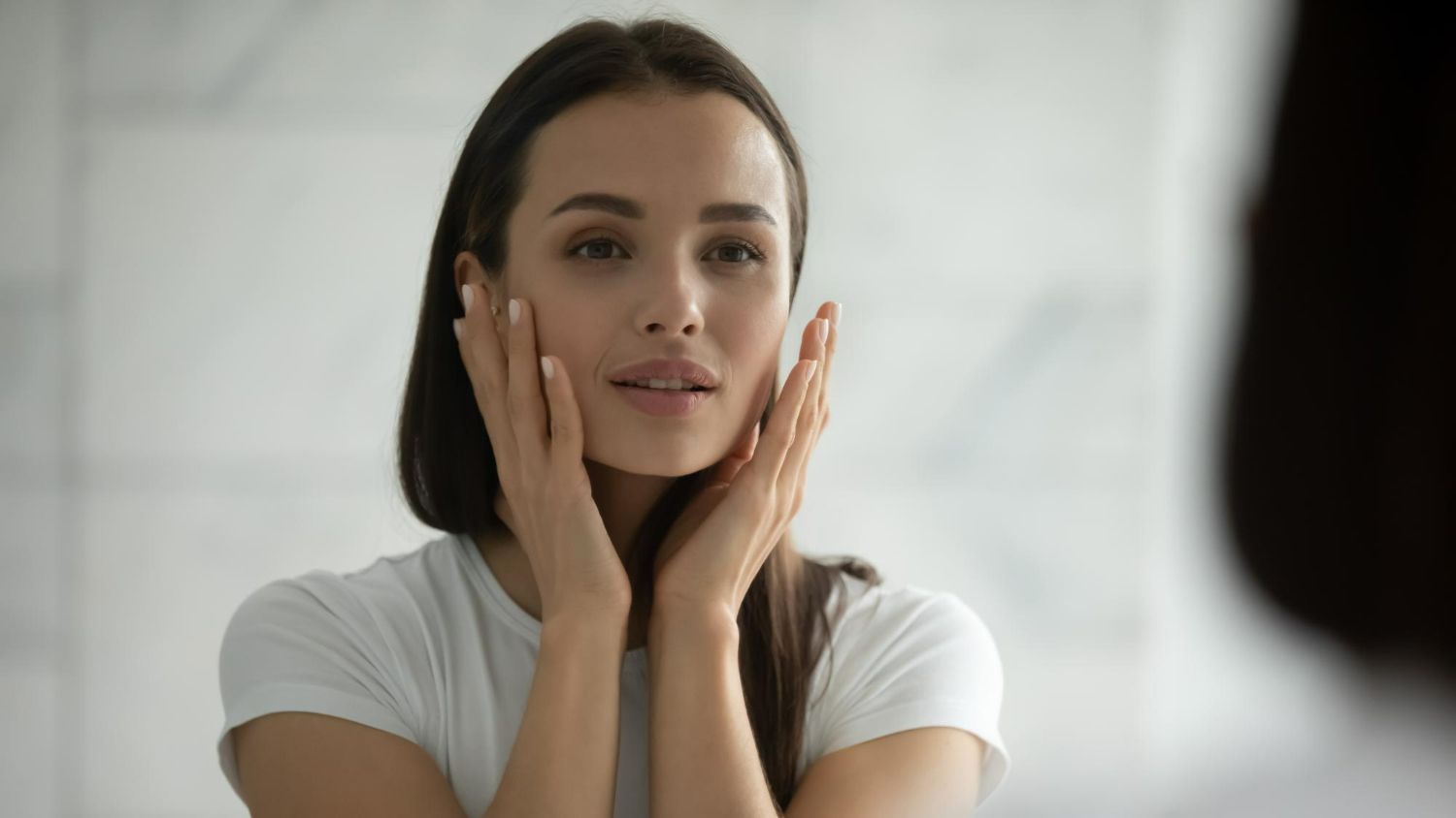Woman in white t-shirt looking in a mirror, touching her face with both hands, in a bathroom.