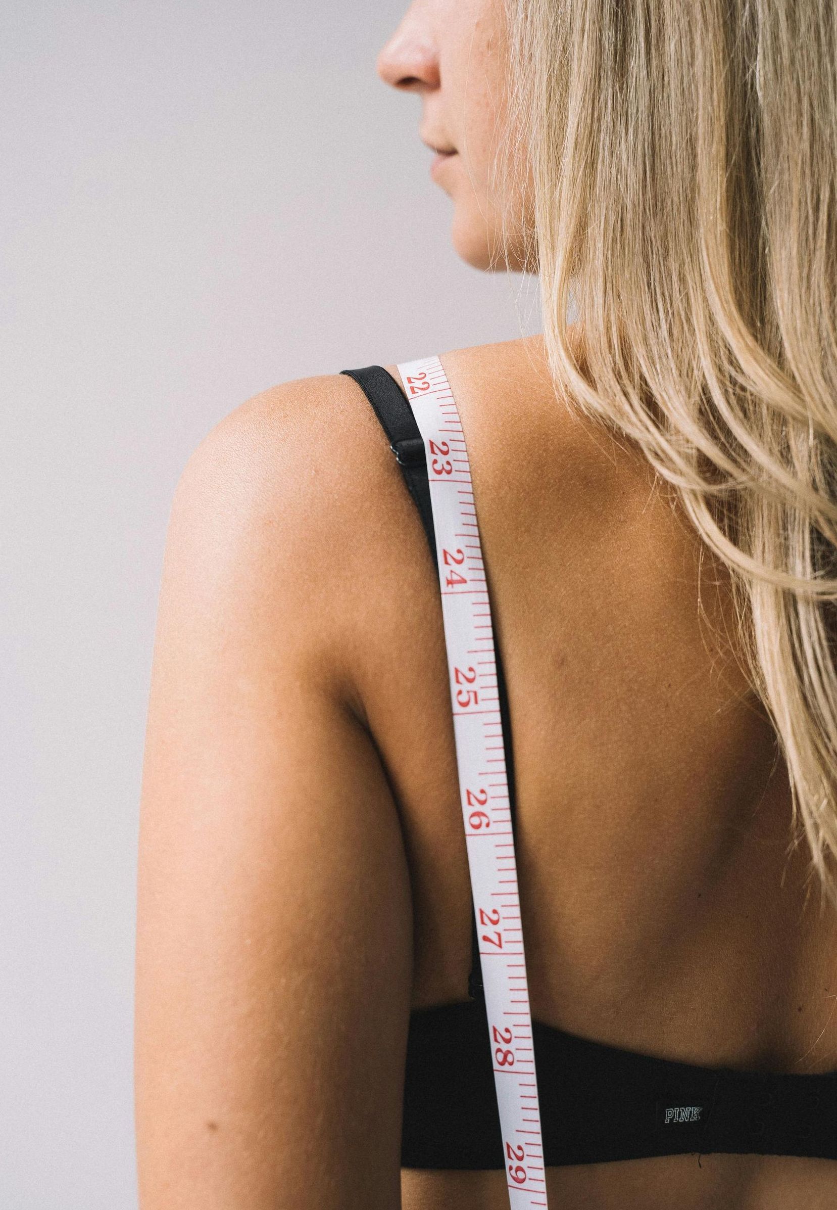 Woman's back and shoulder with a tape measure hanging down, checking a measurement. Blonde hair, neutral background.