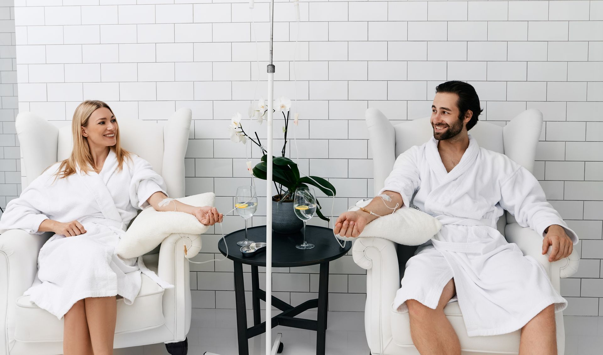 A smiling person and another person, in white robes, sit in armchairs receiving IV drips in a white room.