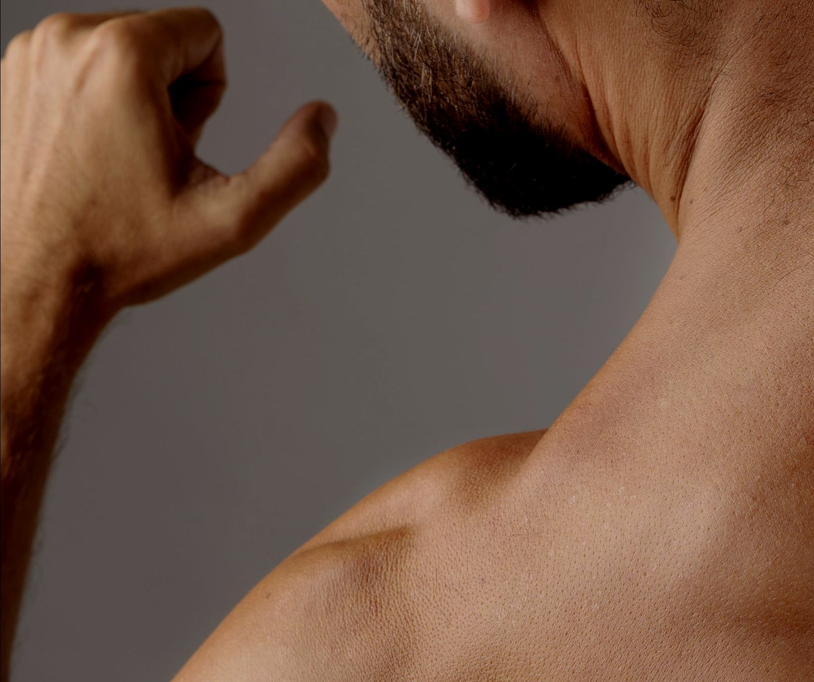 Man flexing bicep, arm raised, tanned skin, gray background.
