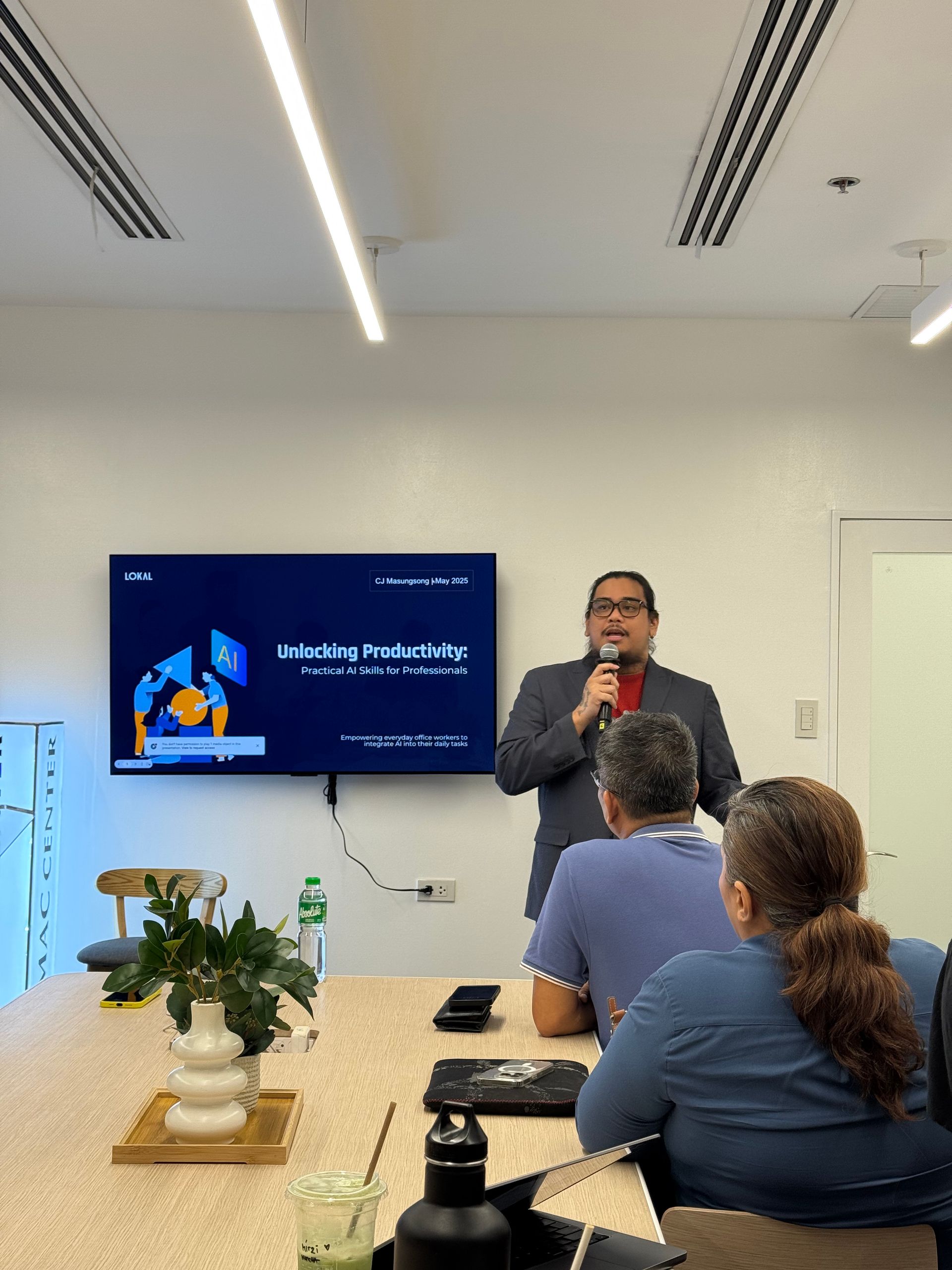 Man speaking at a presentation, screen behind him, audience seated at a conference table.