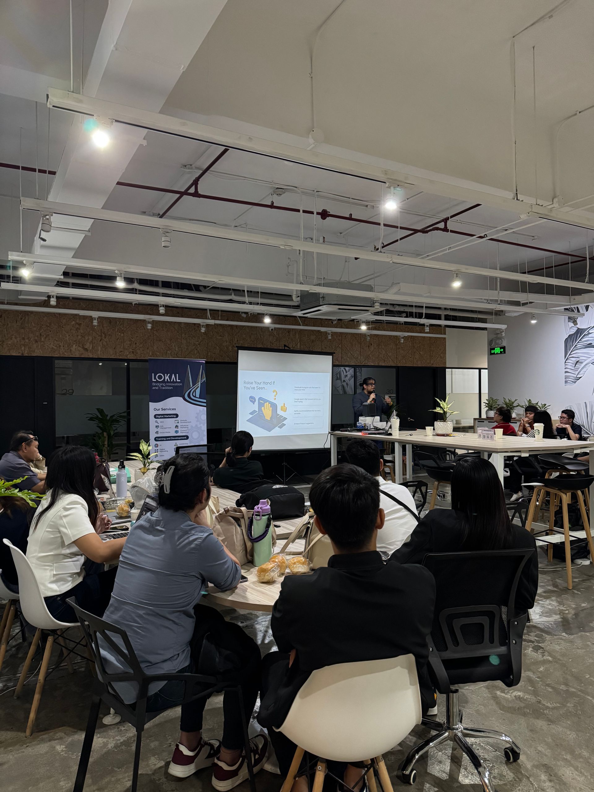 People attending a presentation in a modern office space. A presenter stands near a screen with a graphic.