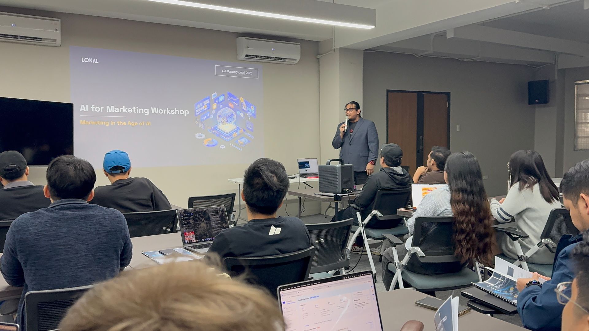 A man lectures in a classroom. Students sit at tables, some with laptops. A projection screen displays a graphic.
