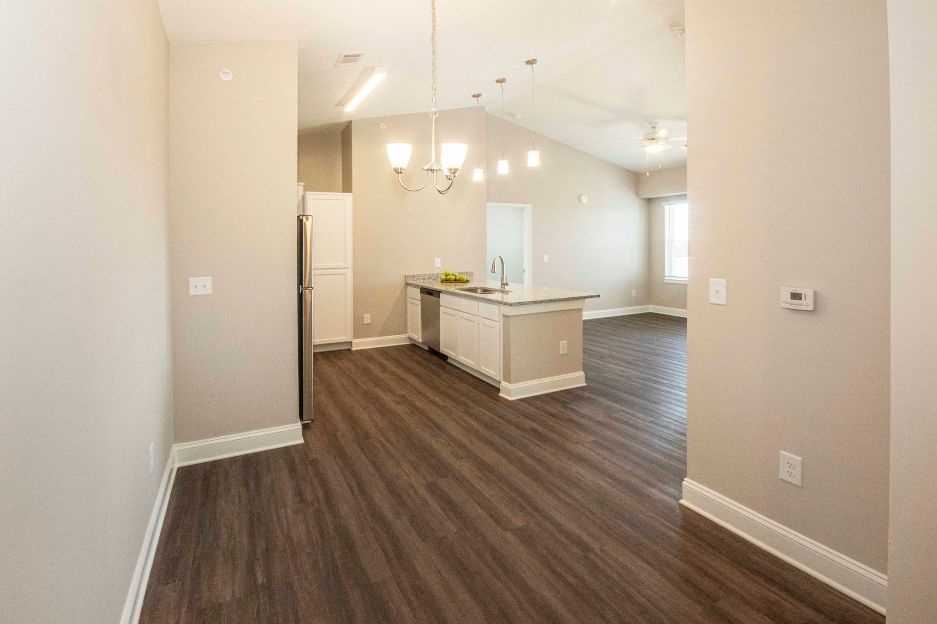 An empty room with hardwood floors and a kitchen in the background.