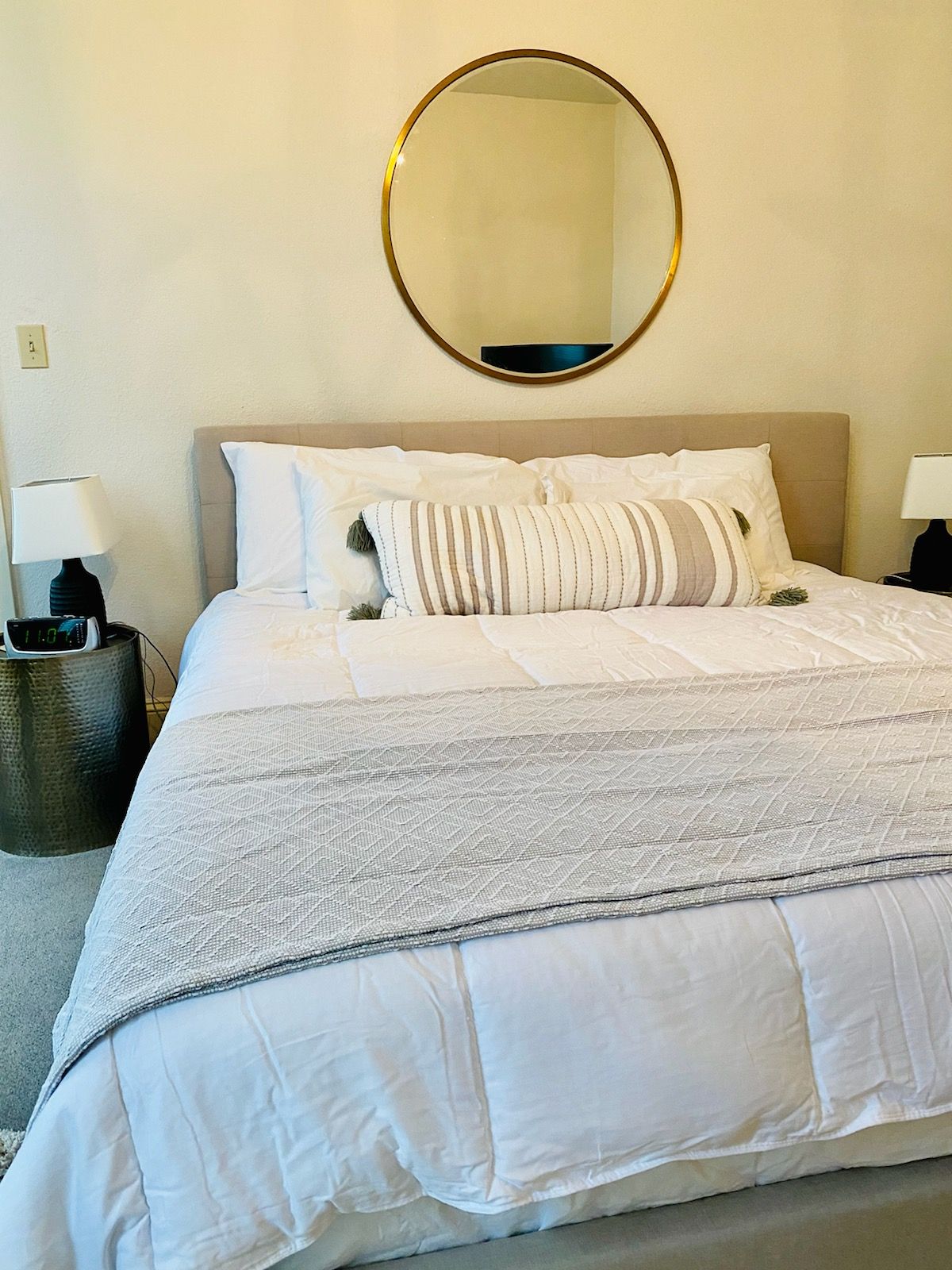 A bed with white sheets and pillows in a bedroom with a round mirror on the wall.