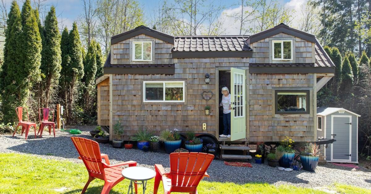 An woman standing in her tiny home's entryway, holding open the front door. Two lawn chairs sit next to a table in the yard.