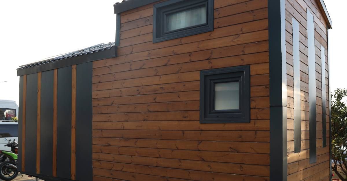 A tiny mobile home made out of brown wooden slats and parked on wheels. The roof and accents are black.
