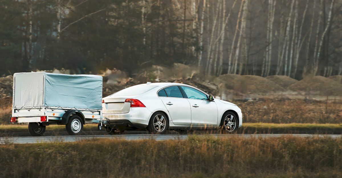 A white sedan drives on a road while pulling a small, two-wheeled trailer that is enclosed by a gray cover.