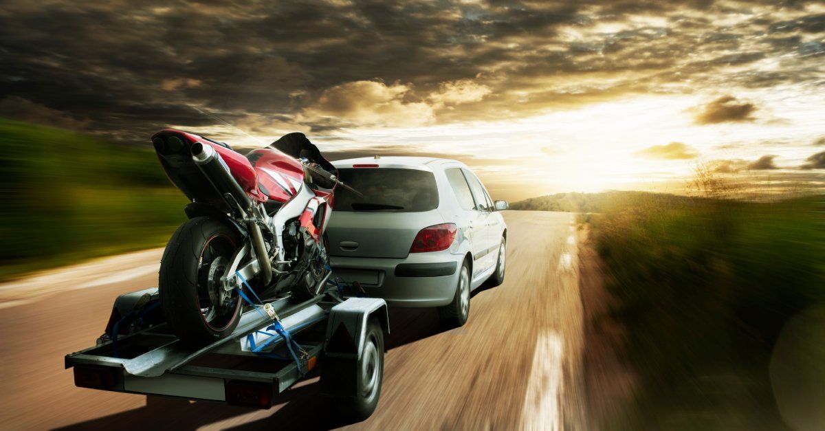 A red motorcycle attached to a two-wheeled trailer pulled by a white minivan on a long winding road.