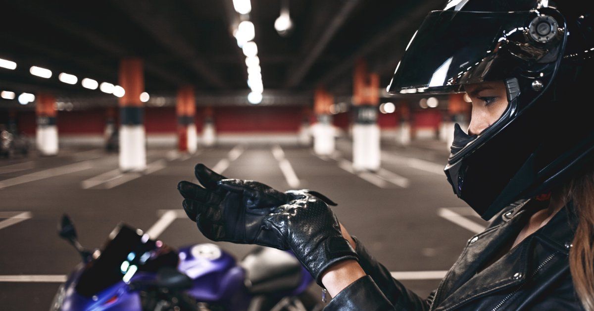 A woman wearing a protective black helmet pulls on a pair of black gloves as she stands near a motorcycle.