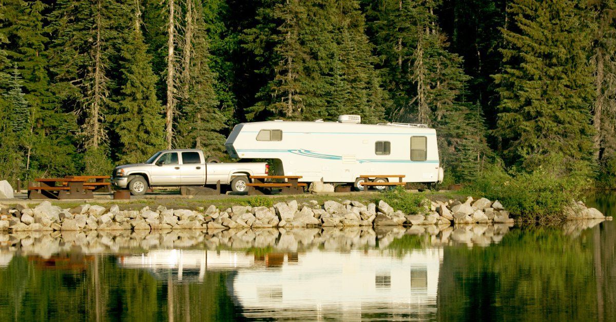 A silver pickup truck with a large white caravan attached to it is parked at a campsite near a lake.
