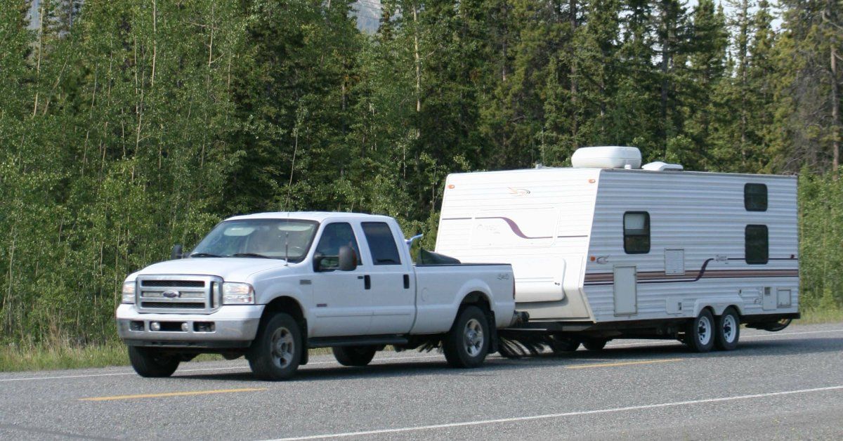 A white pickup truck tows a large white caravan down a road with trees and a mountain in the distance.