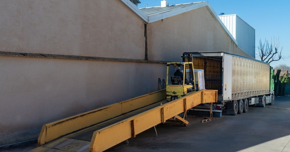 A yellow forklift moves plastic-wrapped pallets up a mobile ramp toward a delivery truck by a beige building.