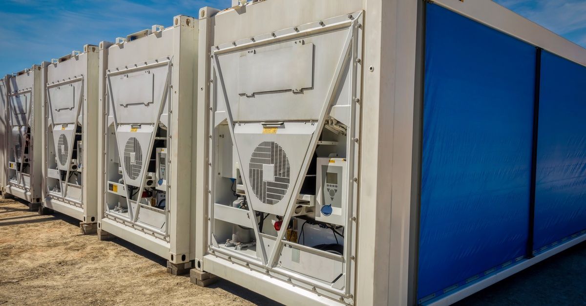Four white refrigerated containers sit on gray bricks, with blue fabric covering the doors of the end unit.