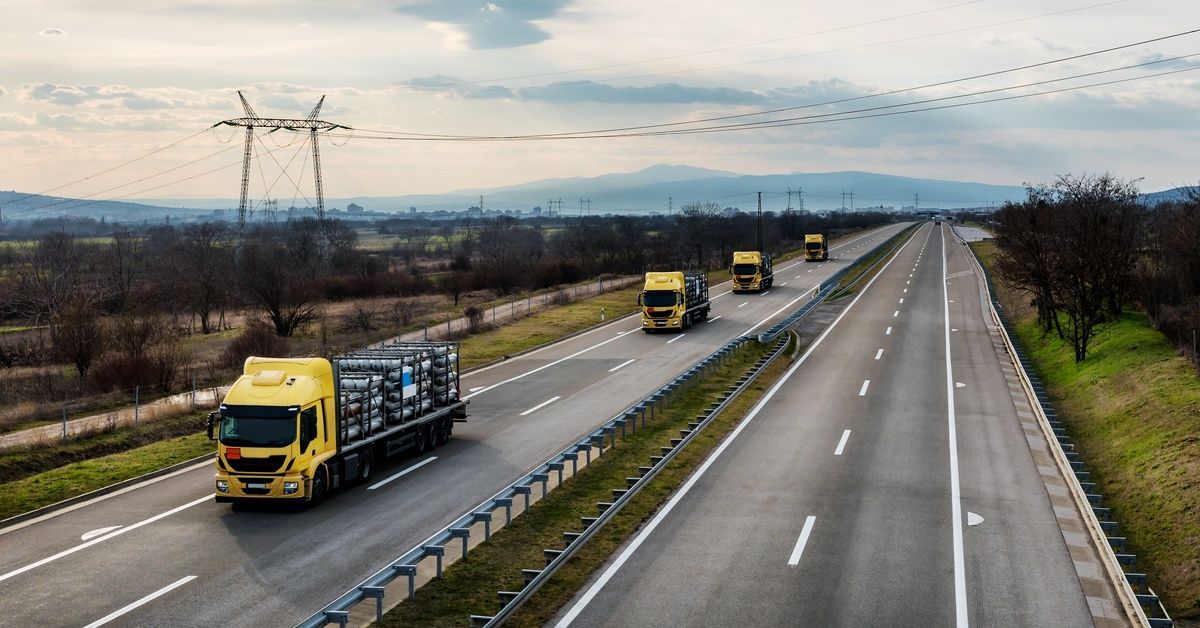 Four yellow trucks carry silver gas canisters on flatbeds while driving in one lane of a four-lane h
