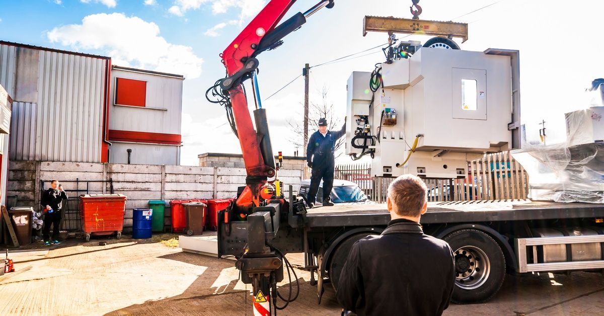 A crane lifts a heavy machine onto the back of a semi. Three men supervise the transfer of this equipment.