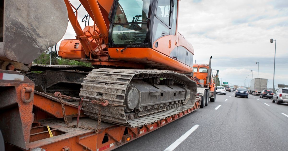 A side view of a semi-truck driving down a highway with an oversized construction machine. Other car