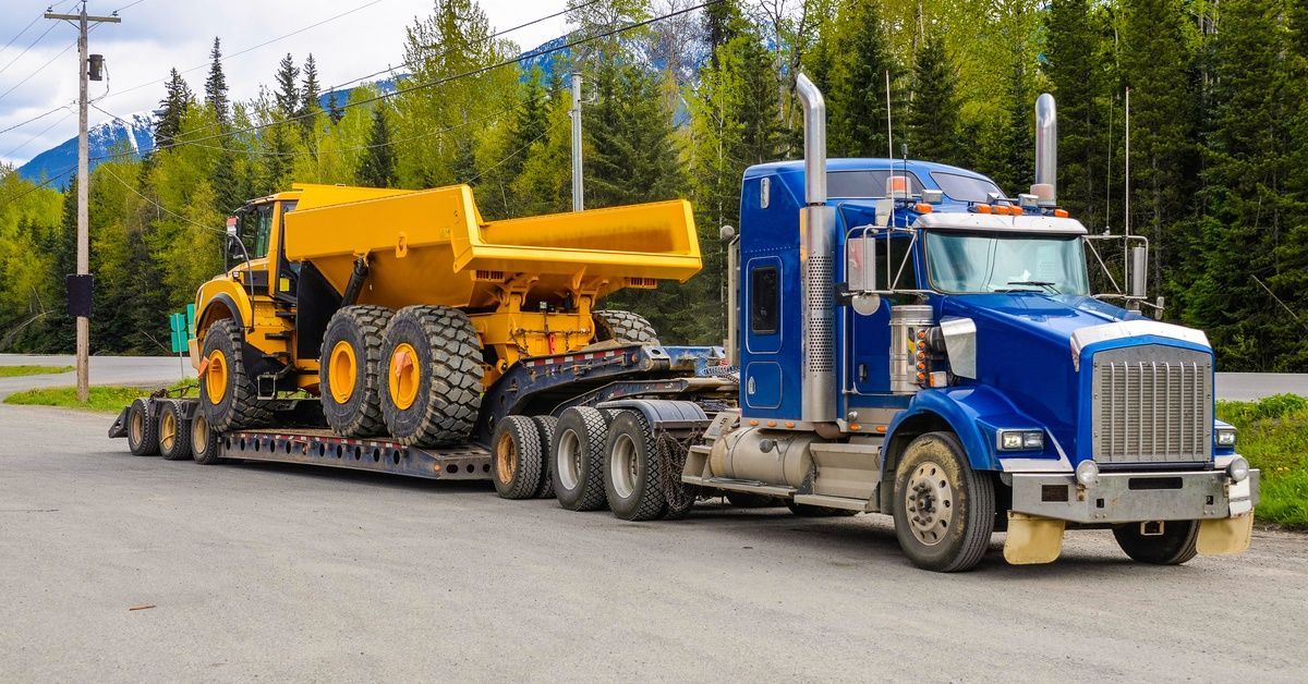A semi parked in a gravel lot with trees in the background. A large construction truck is secured to the back of the semi.