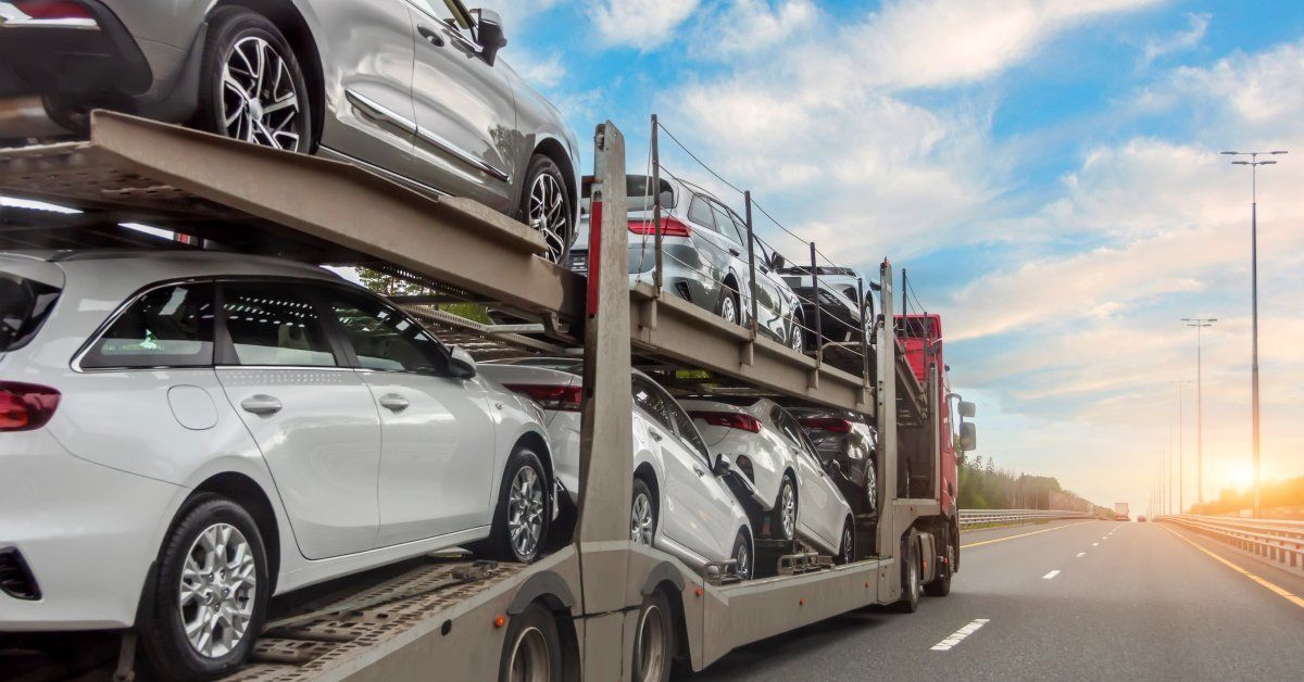 A side view of a semi transporting multiple vehicles. The semi is driving on a road with a blue, cloudy sky above.
