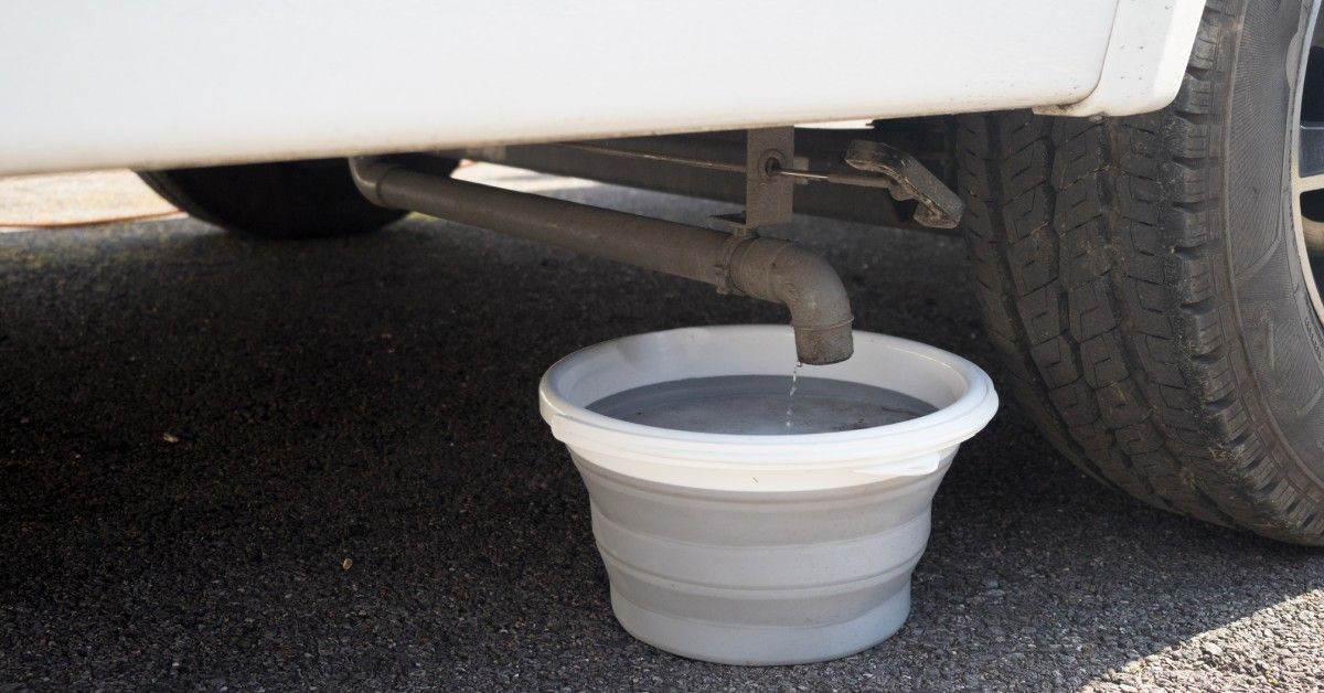 A small collapsible bucket rests under a waste pipe on an RV. The bucket is full as water slowly drips out of the pipe.