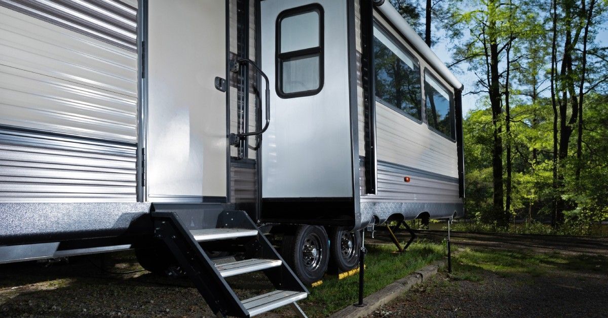 The side of an RV with a slide-out and stairs pulled out. There are trees and a blue sky in the background.