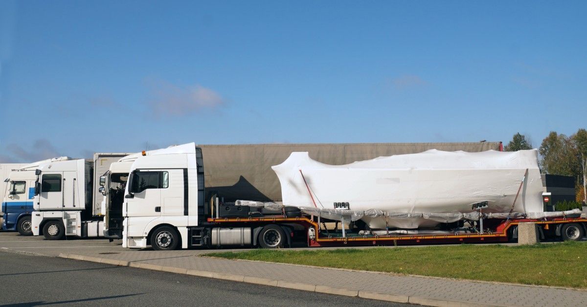 A few semi trucks are parked next to each other. The front semi has a large boat shrink-wrapped on its trailer.