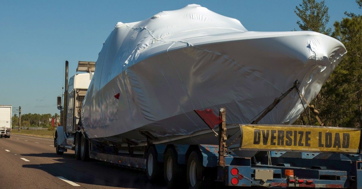 A yacht in white shrink wrap rests on a trailer attached to a semi. The semi drives on a highway.