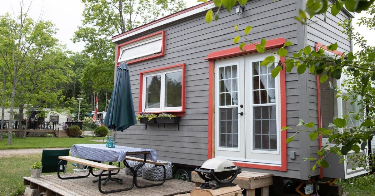A tiny home with grey siding and red trim. There is also a deck with a picnic table and portable grill in front of the home.