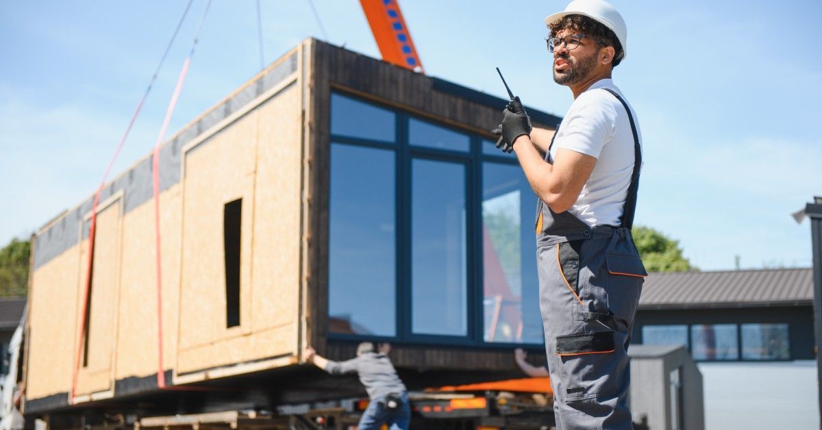 A professional mover holds a walkie-talkie and wears a hard hat. Movers position a tiny home on a trailer in the background.
