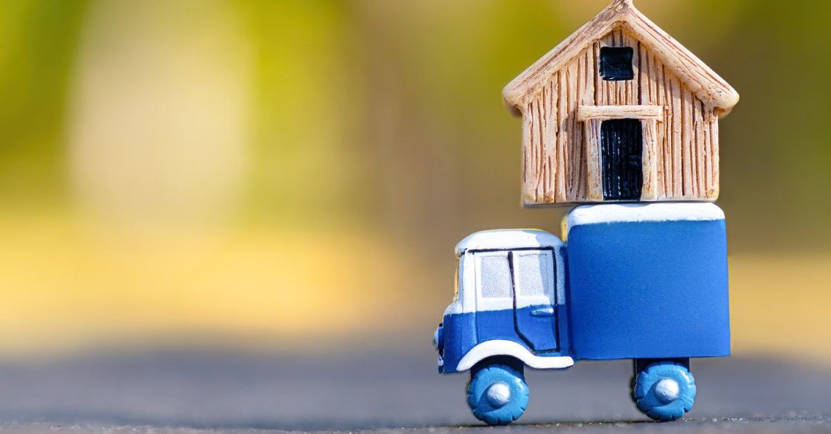 A blue toy truck with oversized wheels carries a small wooden house on its bed.