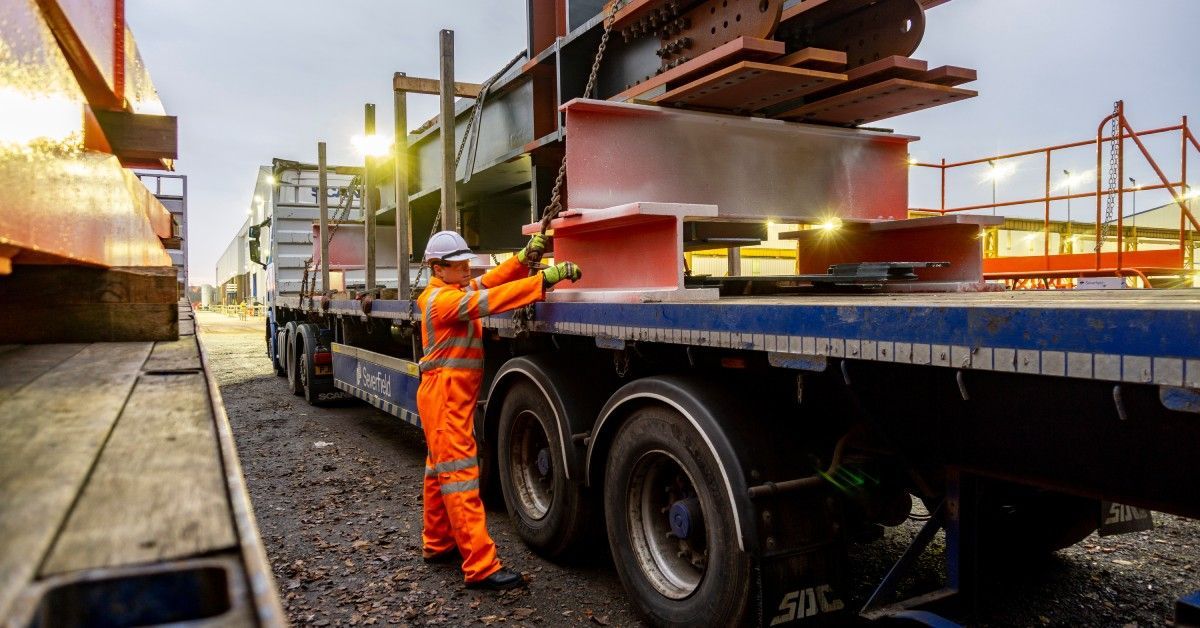 A person wearing an orange safety suit and a white hard hat loads a flatbed trailer with heavy, metal beams.