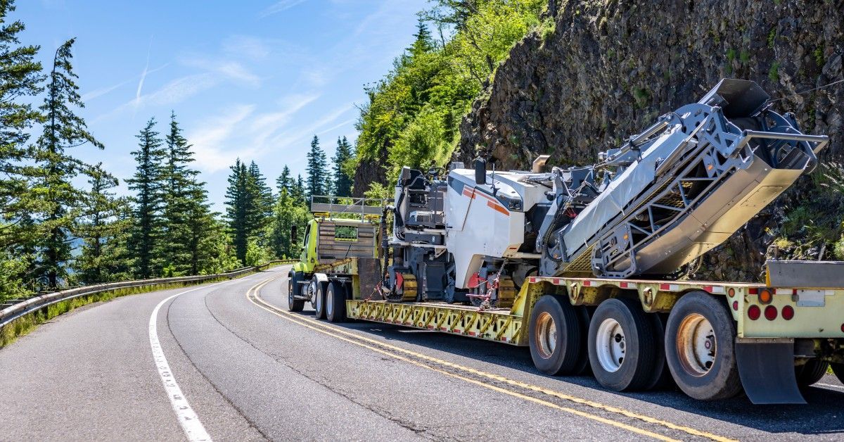 A trailer truck carrying a semi rig drives through a windy road. Trees are on the left side of the road with a cliff on the right.