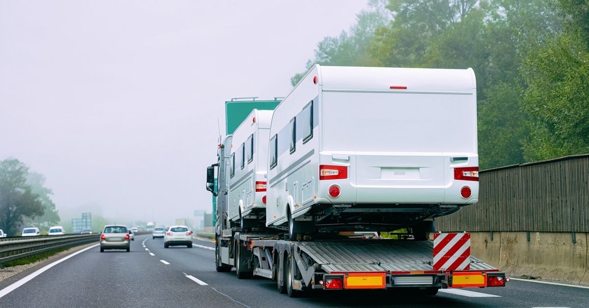 Two campers are strapped to a trailer attached to a semi truck. The truck drives down a highway with cars ahead of it.