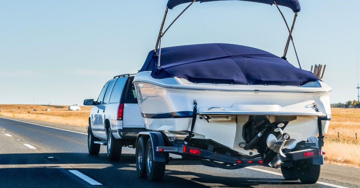 A truck tows a boat on a trailer down a road. Dry, dying grass lines the road to the right of the truck.