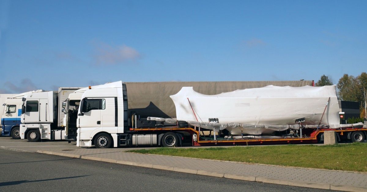 A semi truck with a large boat covered in white shrink wrap on the back of the trailer. Other semis are parked nearby.