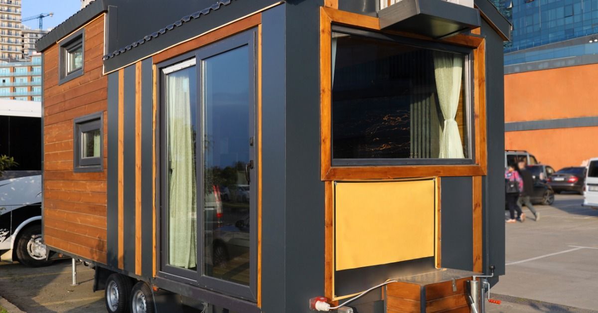 A tiny home on a trailer is parked in a parking lot. The home has wood siding with black accents.