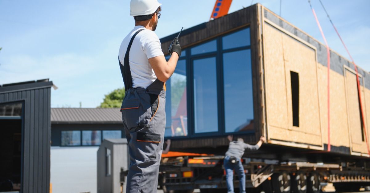 Someone wearing a white hard hat and holding a walkie-talkie supervises a tiny home being placed onto a trailer.
