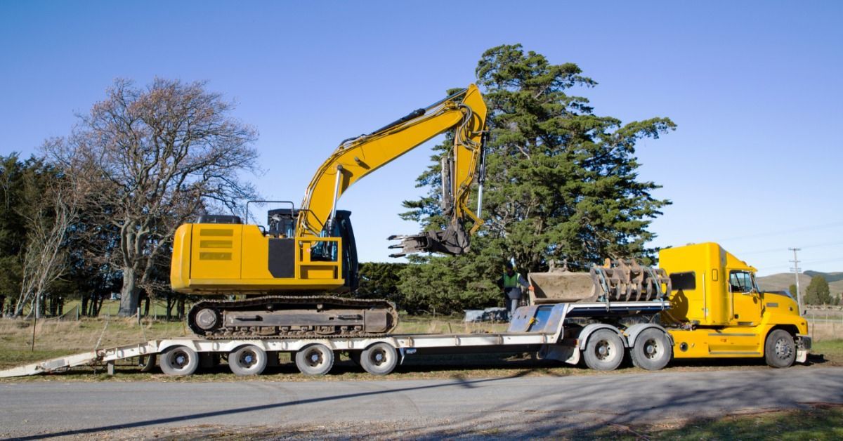 A semi with a long flatbed trailer parked in some grass next to a driveway. A large, yellow digger is on top of the trailer.