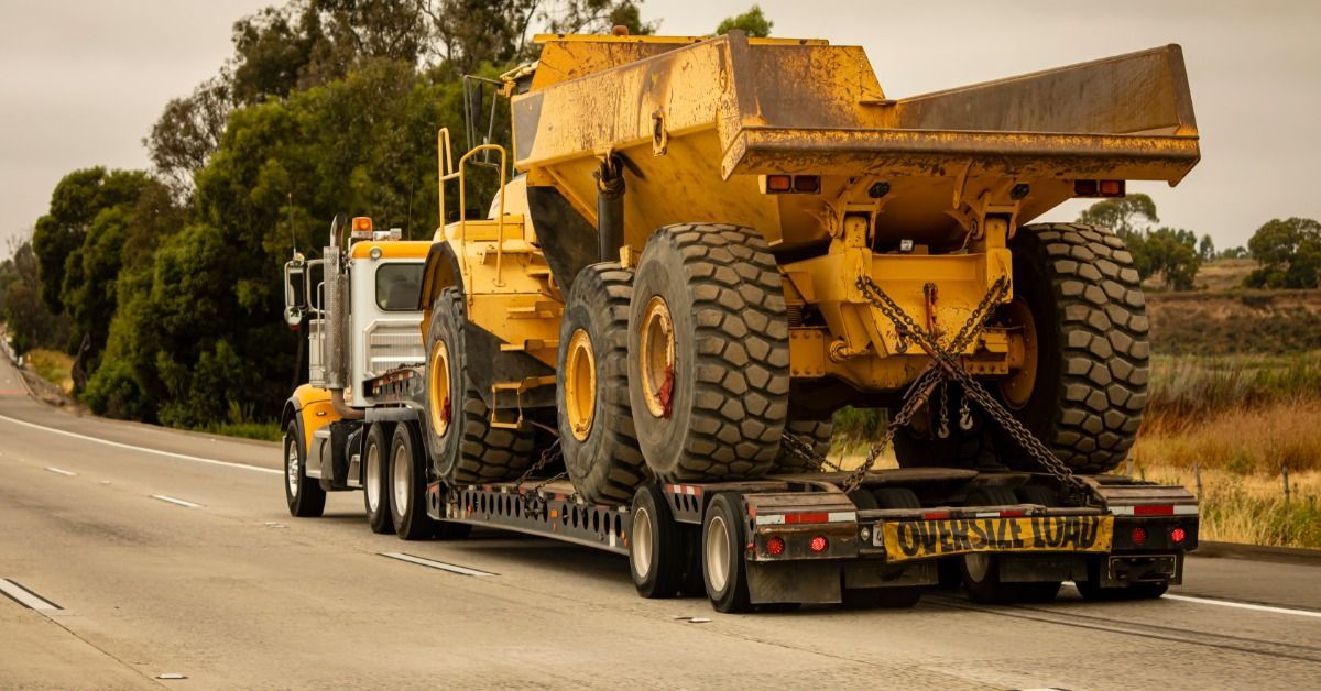 A dirty yellow dump truck strapped to the back of a semi-trailer. The semi drives down a multi-lane road.