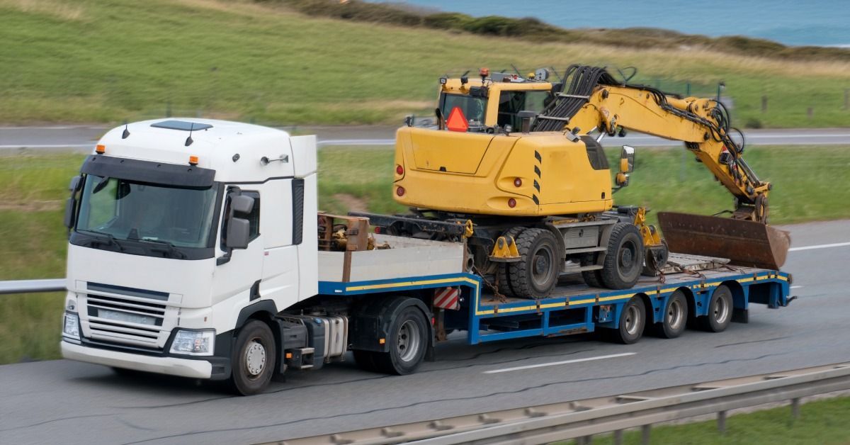 A yellow construction tractor tied down to the back of a semi-trailer. The load drives down a two-lane highway.
