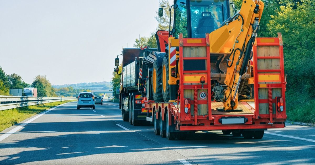 A white semi drives down a road with a large yellow excavator tied down in the back of the trailer.