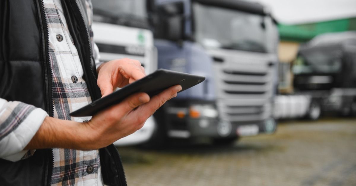 A close-up of someone holding a black tablet in one hand. A few semi trucks are blurred in the back.